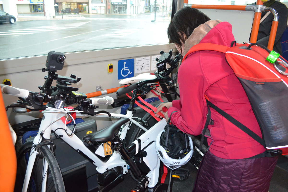 バスの車椅子エリアに自転車を載せ、指定のベルトで固定する>